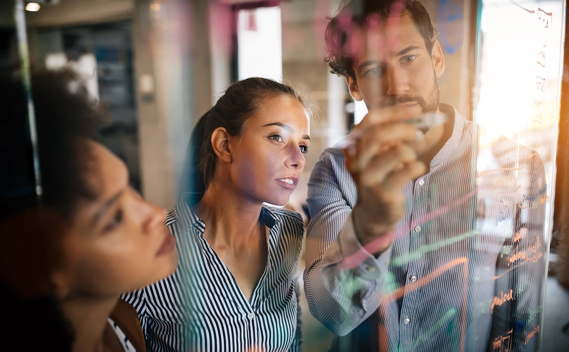 three people, standing in front of transparent screen, one person writing