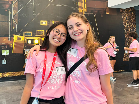 Two women in pink shirts stand in front of a mural picturing a caterpillar truck.