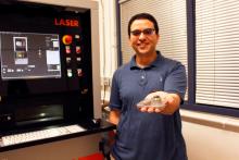 Mohammed Shafae holds out a 3D-printed metal part and stands in front of a 3D printer