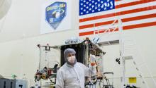 A man in a white cleanroom suit stands in front of a spacecraft under construction, pointing backwards at it. On the wall in the background are a sign for the OSIRIS-REx mission and a large U.S. flag.