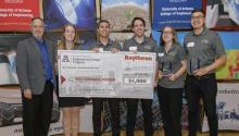 A group of five students in matching grey polos and a man in a blazer hold up a giant, $5,000 check, made out to "Best Overall Design."