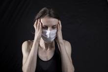 A woman wearing a surgical masks holds her head in her hands and winces, indicating she has a headache.