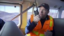 A man wearing an orange safety vest sits inside a van and speaks into a radio handset.