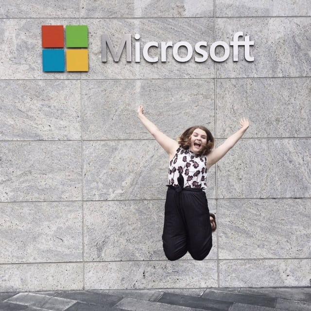 A woman leaps into the air before a gray wall emblazoned with the Microsoft logo.