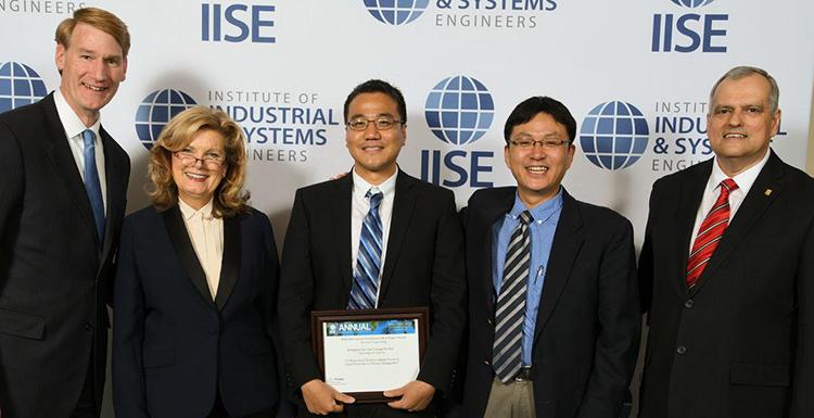 A group of men and a woman holding an award certificate as they stand before a wall with IISE written on it. 
