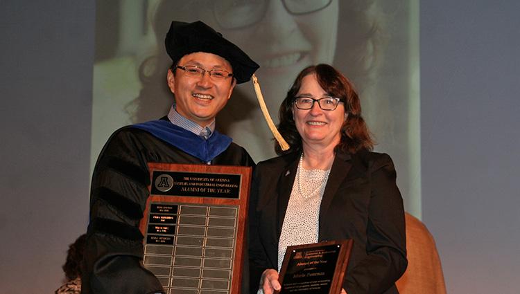 A woman in a black suit jacket accepts a plaque from a man wearing graduation regalia. He is also holding a larger plaque with four names inscribed.