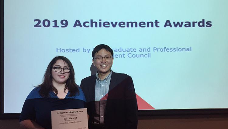 Sara Masoud and Young-Jun Son stand in front of a display screen that reads "2019 Achievement Awards." Sara is holding a certificate with her name on it.