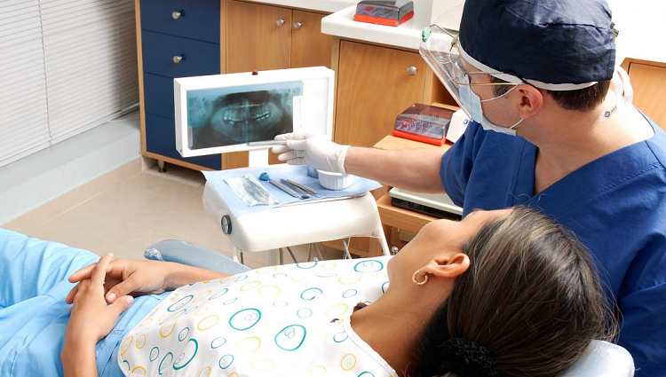 A woman lies in a dentist's chair while a dentist sits next to her wearing a face shield and pointing to an X-ray of teeth.